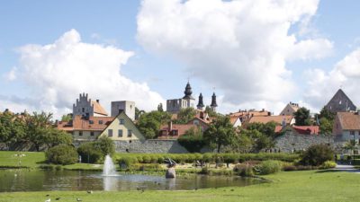 Staden Visby med hus och kyrktorn. Framför staden är en sjö med en liten fontän i.