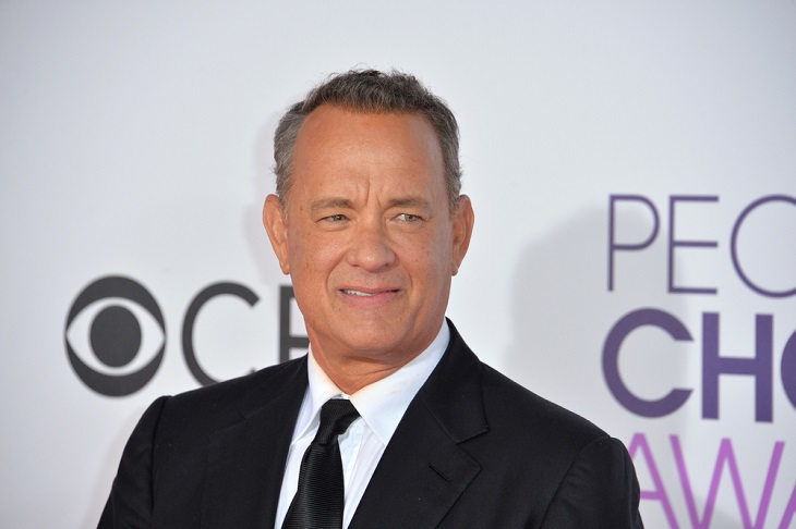 Tom Hanks står fram en vit vägg. Han har kort hår som är lite grått på sidorna. Han har kostym och slips på sig.