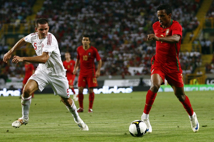 En fotbollsspelar i rött springer med bollen. Bredvid honom springer en spelar i vitt och försöker jaga ikapp honom. Bakom dem syns en stor fotbollsplan och en stor läktare.