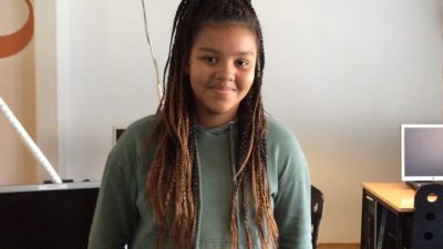 Miriam står och lutar sig mot en skolbänk. Hon har en grön hoodie och långa rastaflätor. Hon ler mot kameran.