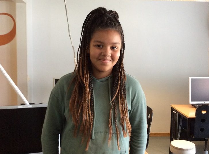 Miriam står och lutar sig mot en skolbänk. Hon har en grön hoodie och långa rastaflätor. Hon ler mot kameran.