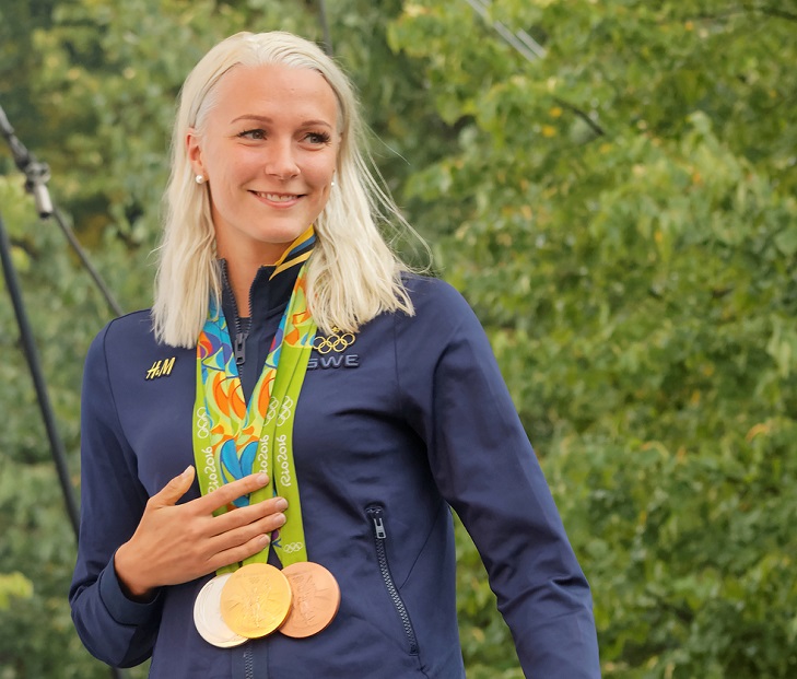 Sara Sjöström. Bakom henne syns massa gröna löv. Hon har långt ljust hår och en blå jacka. Runt halsen hänger flera medaljer i band.