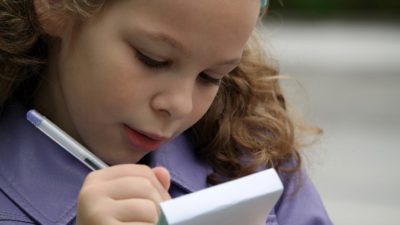 En flicka skriver i ett block. Hon har lockigt blont hår och en lila jacka.