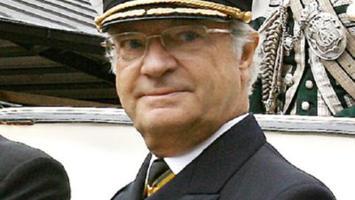 Kungen tittar snett bortåt. Han har en hatt med skärm och guldiga symboler. Han har glasögon och vitt hår. Han har en svart kostymjacka med flera medaljer på bröstet.