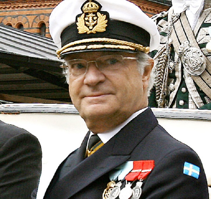 Kungen tittar snett bortåt. Han har en hatt med skärm och guldiga symboler. Han har glasögon och vitt hår. Han har en svart kostymjacka med flera medaljer på bröstet.