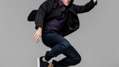 Benjamin hoppar upp i luften. Han har blå jeans, rutig skjorta och en svart jacka på sig.