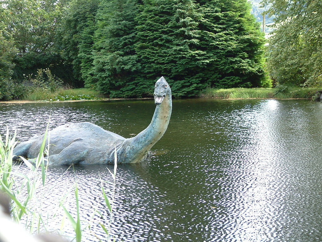 En sjö med träd vid sjökanten. I sjön ligger en stor staty. Statyn har avlång men stor kropp, lång halv, litet huvud och fenor. Den öppnar munnen och gapar mot oss.