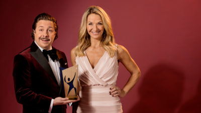 David och Kristin står i finkläder och ler stort. David håller i ett av galans priser. Priset är av metall och föreställer en siluett av en figur som höjer armarna.