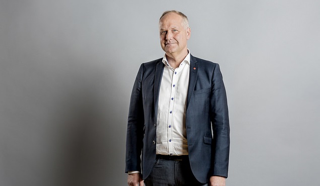 Jonas står framför en vit vägg. Han har kostym och skjorta på sig. Han har kort grått hår och ser allvarlig ut.